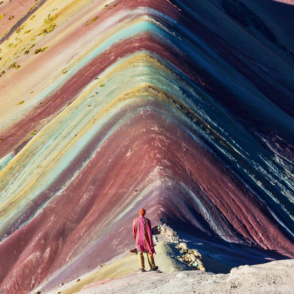 Traveler standing at the summit ridge of Rainbow Mountain overlooking the vibrant natural colors of Vinicunca in Peru.