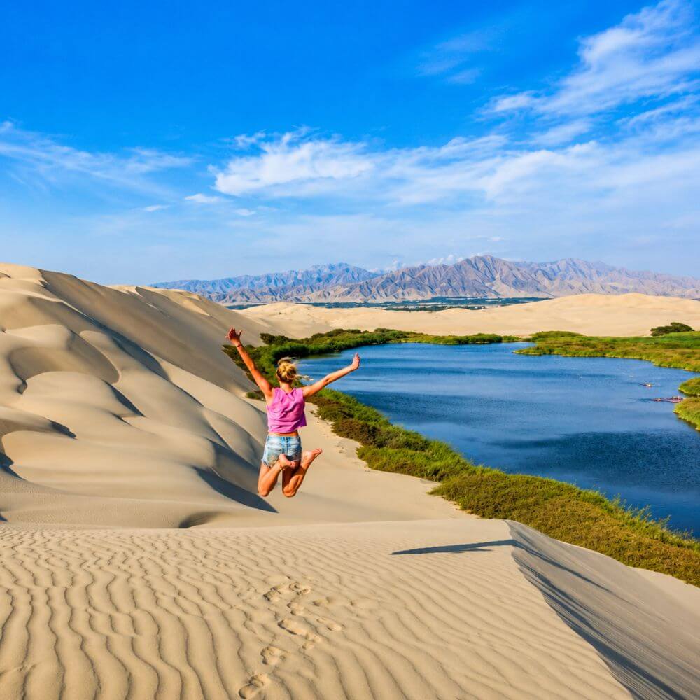 Panoramic view of Oasis Morón with golden sand dunes, blue lagoon, and lush vegetation near Paracas, Peru.