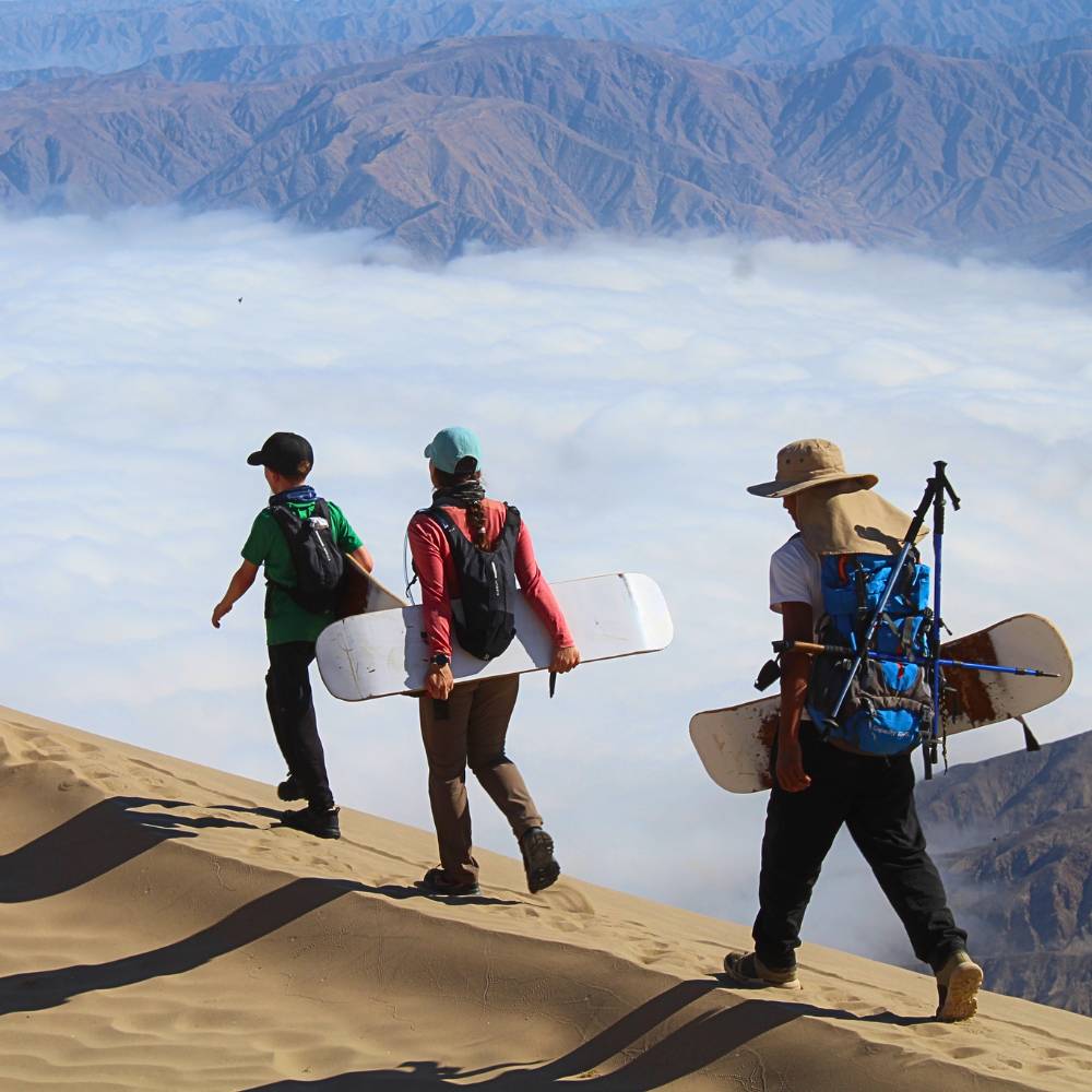 Adventure travelers carrying sandboards across the giant dunes of Cerro Blanco in the Nazca Desert.