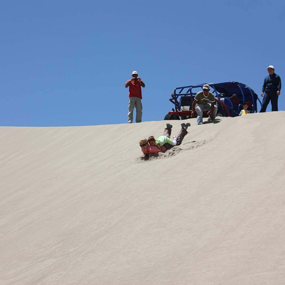 Travelers enjoying sandboarding on the towering dunes of the Usaka Desert during an exciting USAKA DESERT TOUR in Nazca, Peru.