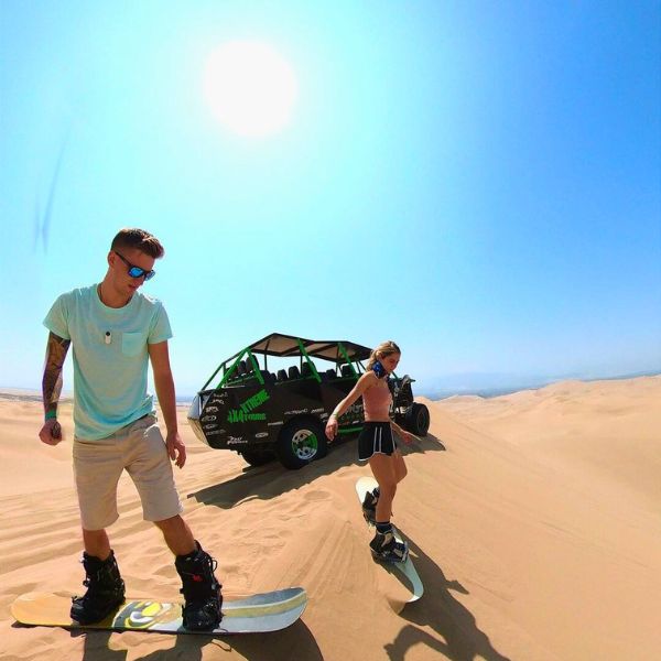 Travelers sandboarding in the Huacachina desert with dune buggy nearby