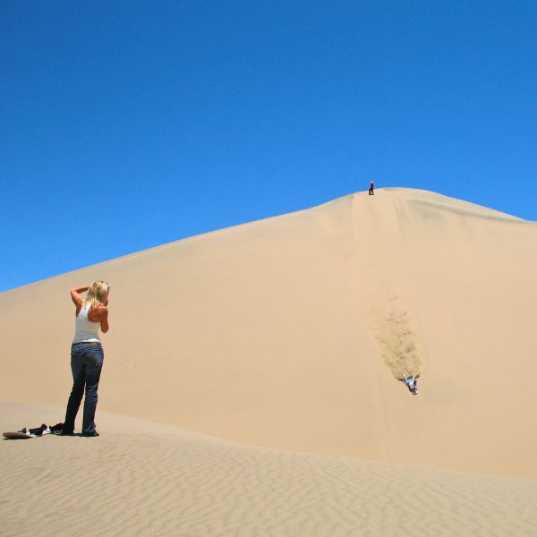 Visitor photographing sandboarder on a steep dune in Usaka Desert